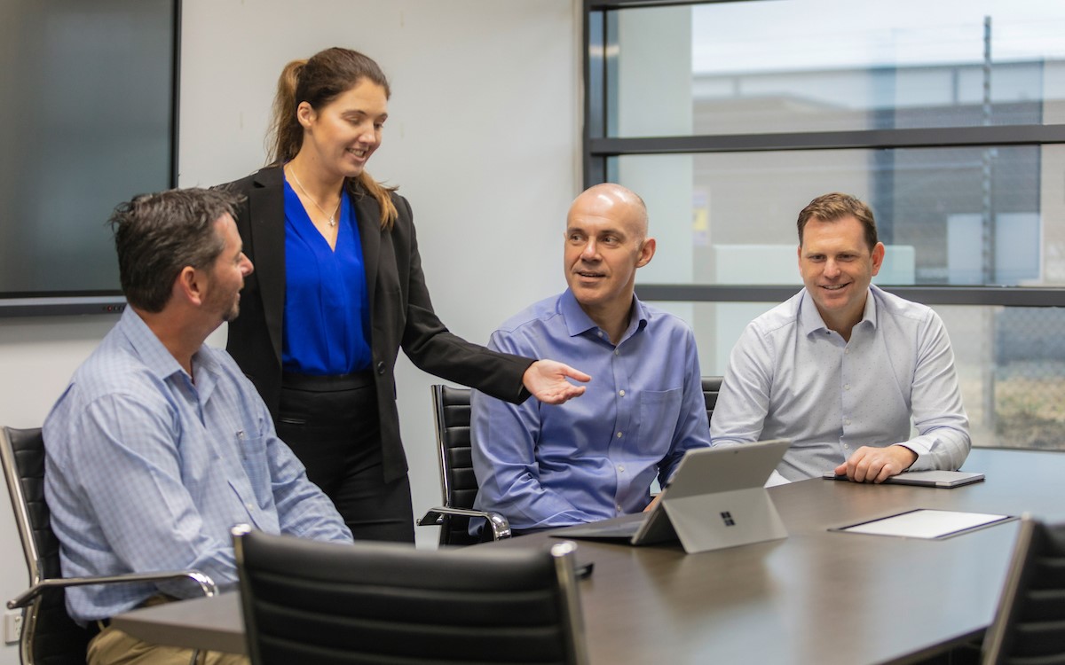 A woman standing and three men sitting around a laptop