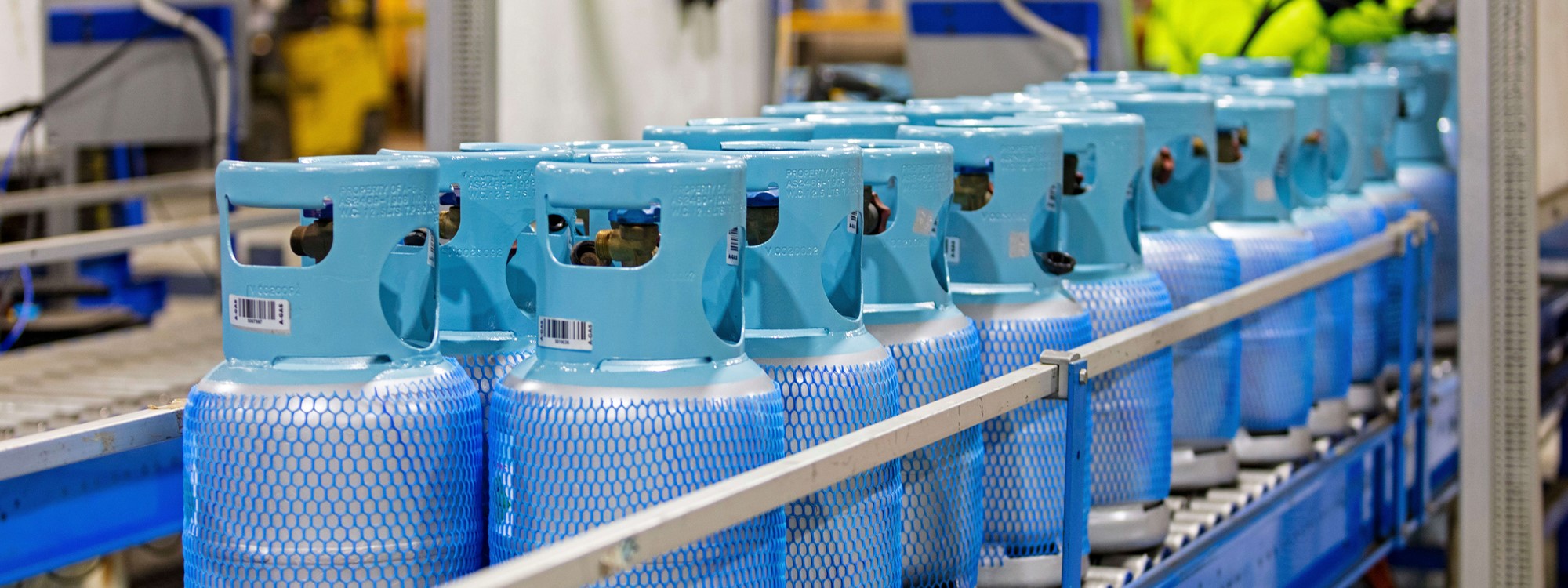 A row of grey and blue cylinders wrapped in blue casing on a shelf with an operator in the background 